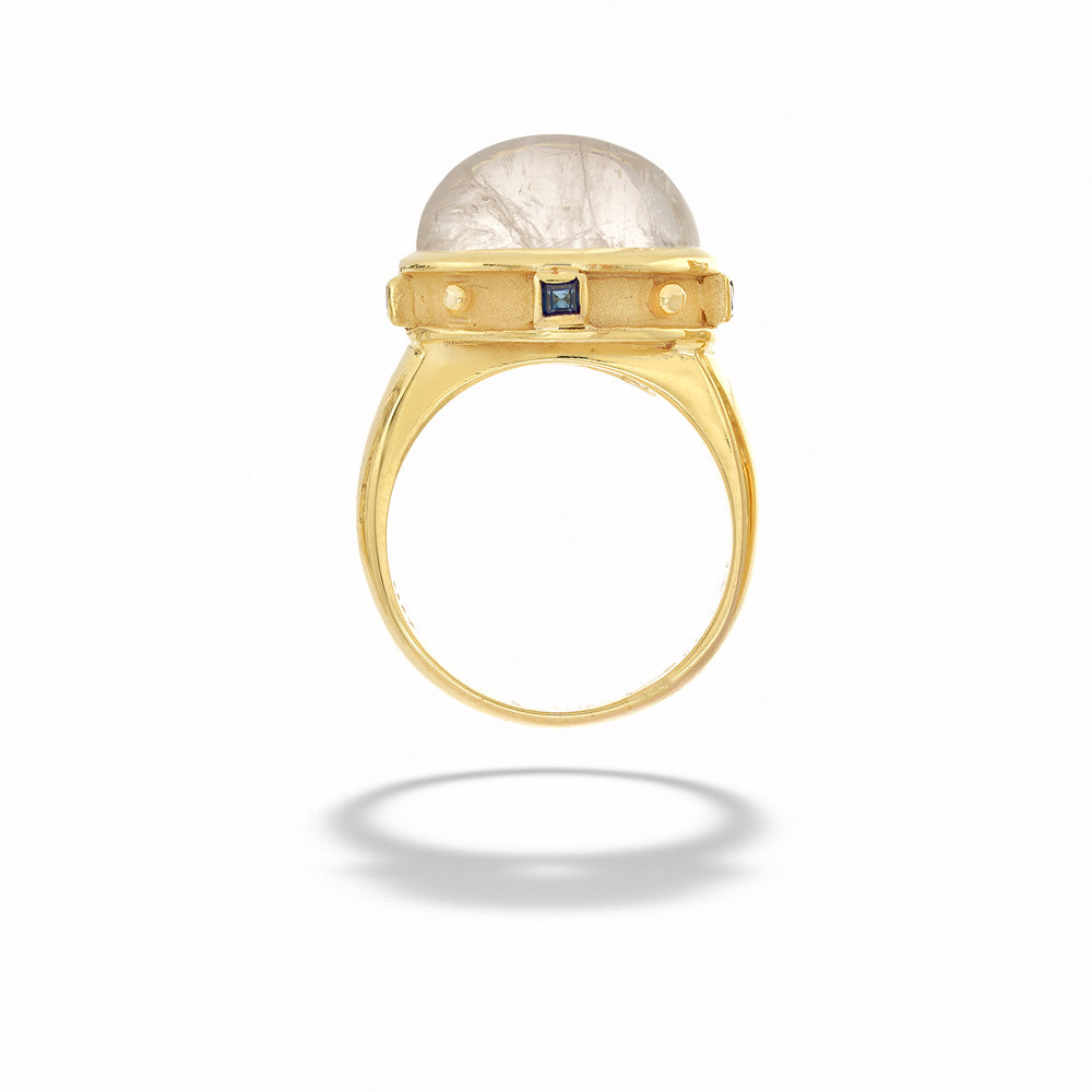 Gold ring with a large central stone and smaller stones on a white background Moonstone Ring with Faceted Blue Sapphires by Mazza