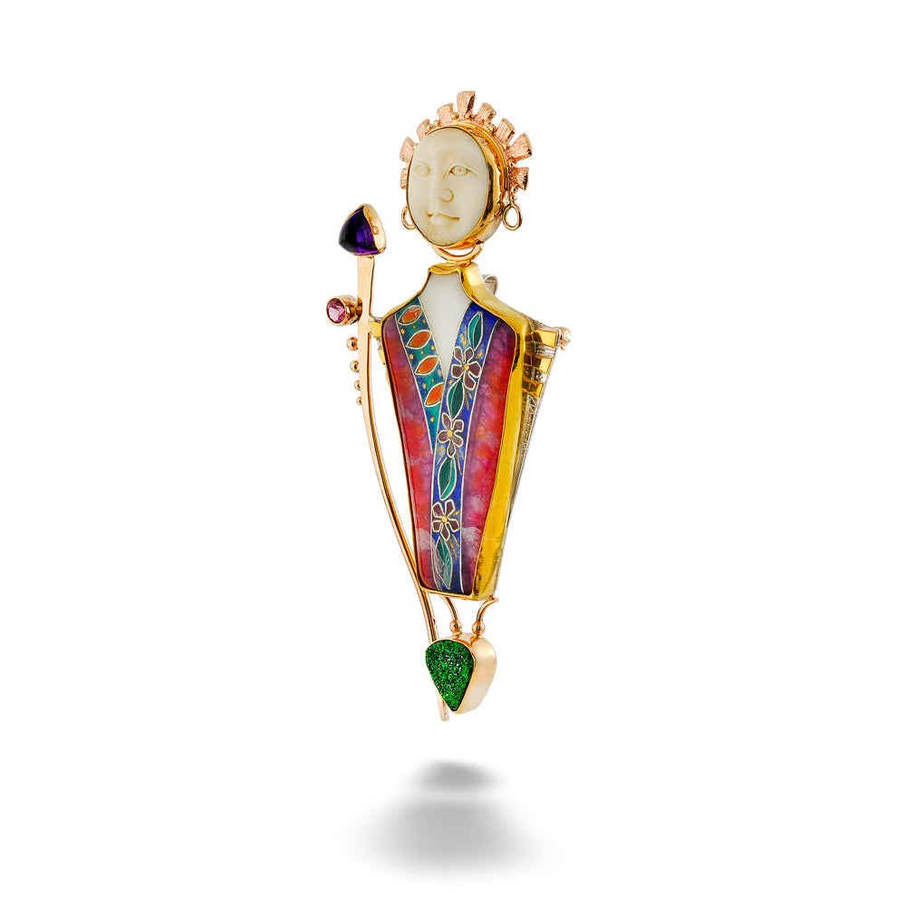 A gold-toned pendant brooch shaped like a stylized figure with a carved face, holding a staff with a black and purple top, decorated with colorful enamel panels in pink, blue, green, and gold, set against a plain white background. The Goddess Pendant by Katrin Wolf