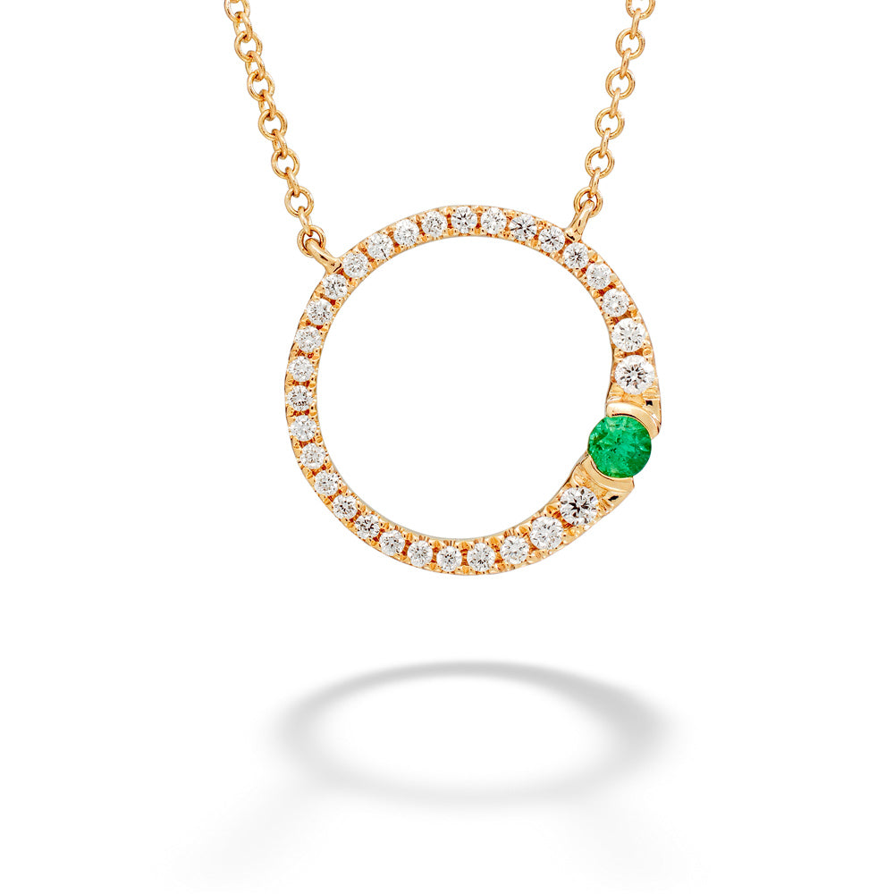 A gold necklace with a crescent-shaped open pendant, paved with small clear stones and featuring a single round green emerald on the right side, shown against a white background.
